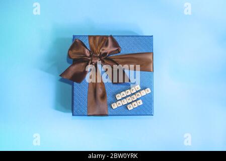 Ein blaues Geschenk mit braunem Streifen auf dem blauen Hintergrund. Ein Schild Happy Father's Day aus Holzblöcken. Konzept des Vatertags. Stockfoto