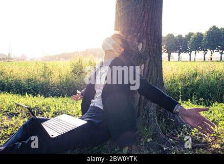 Junger, tausendjähriger lächelnder Junge, der am Computer arbeitet, entspannt unter einer Pflanze an einem sonnigen Tag im Freien sitzt - Konzept über Smartwork in der Natur mit CO Stockfoto