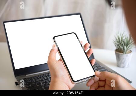 Mockup Hand hält leere weißen Bildschirm Smartphone mit leeren weißen Laptop-Bildschirm auf Home Office Arbeitstisch. Mock-up Smartphone und Computer-Bildschirm Stockfoto