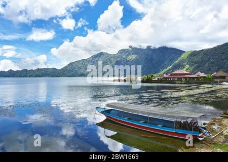 Ein leeres Touristenboot, das sich am Rande des Batur Sees vor einer Kulisse von Bergen und einem wunderschönen, wolkigen blauen Himmel lehnt. Kintamani, Bali, Indonesien Stockfoto