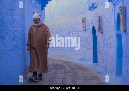 Ein einheimischer Marokkaner in traditioneller Kleidung geht eine schmale, gepflasterte Gasse in der Altstadt von Chefchaouen, Marokko, entlang. Stockfoto
