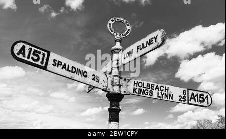 Ein altmodisches britisches Straßenschild auf der A151, das auf Spalding, Holbeach, Kings Lynn und Low Fulney Estate in schwarz und weiß vor Wolken zeigt Stockfoto