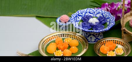 Nahaufnahme mehrerer traditioneller thailändischer Nachspeisen, die auf einem Porzellan- und Messingtablett mit Blumen- und Bananenblättern auf einem weißen Tisch serviert werden Stockfoto