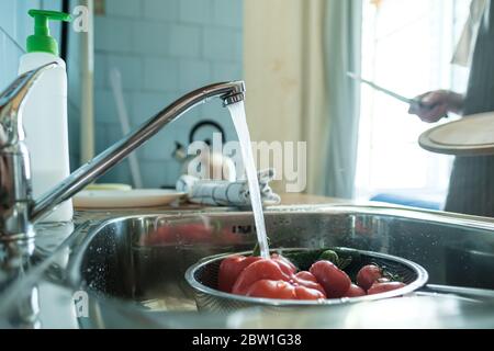 Frisches Gemüse unter einem Wasserstrom in einem Sieb in der Spüle, auf einem Hintergrund weibliche Hand mit einem Messer und einem Schneidebrett. Stockfoto