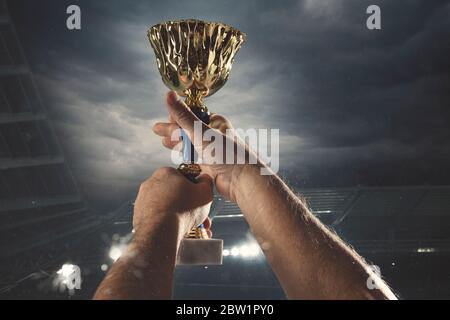 Traum wird wahr. Auszeichnung des Sieges, männliche Hände straffen die goldene Tasse der Gewinner gegen bewölkten dunklen Himmel. Sport, Wettbewerb, Meisterschaft, gewinnen, das Ziel erreichen. Preis für Erfolg und Ehre. Stockfoto
