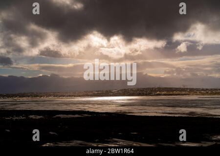 Sonnenstrahlen, die durch die dunklen Wolken scheinen, erhellen einen kleinen See an einem kalten Tag. Berge, die aus der Landschaft in der Ferne steigen. Stockfoto