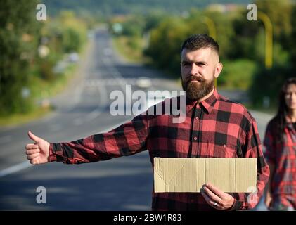 Mann mit strengem Gesicht und Bart, der durch Anhalter mit Straße auf Hintergrund reist. Reise- und Anhalter-Konzept. Hipster mit Mädchen versuchen, Auto mit leerem Karton Zeichen und Daumen nach oben Geste stoppen. Stockfoto