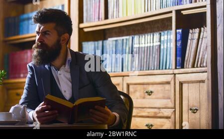 Altmodischer Mann in der Nähe der Teekanne. Aristokratisches Lifestyle-Konzept. Aristokrat auf strikte Gesicht Lesebuch. Mann in klassischen Anzug sitzt im Vintage-Interieur, Bibliothek, Bücherregale im Hintergrund. Stockfoto