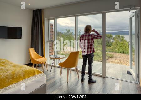 Telearbeit. Mann mittleren Alters mit lockigen blonden Haaren, rot-weiß kariertem Hemd auf dem Rücken und einer Tasse Kaffee nach draußen Stockfoto
