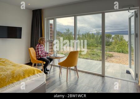 Telearbeit. Mann mittleren Alters mit lockigen blonden Haaren, weiß-rot kariertes Hemd. Mit einem Laptop an einem Tisch in einem modernen Hotelzimmer neben seinem Bett sitzen Stockfoto