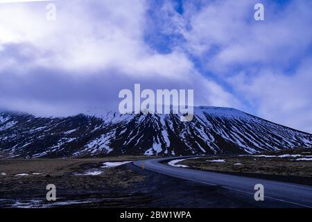 Eine wolkenbedeckte verschneite Bergkette, die teilweise von Wolken verdeckt ist Erhebt sich über eine karge isländische Landschaft mit einer einzigen Straße Durch die Landschaft gewellt Stockfoto