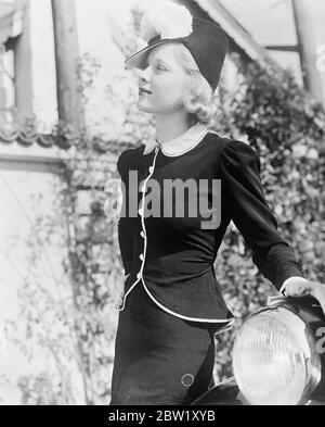 Mary Carlisle, die Hollywood-Filmschauspielerin, trägt ein neues zweiteiliges Modell von Krepp [crêpe/crape] mit einer Jugendlichen Tunika in Weiß mit schmalem Silberrand. Die Knöpfe sind winzige Strass, und eine Schärpe der schwarzen Kreppbinder über den Rücken. Der Hut ist ein smarter schwarzer Stroh mit einer hohen Krone, die von zwei Federbommeln überragt wird. Mai 1937 Stockfoto