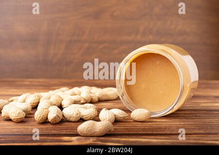 Cremige Erdnusspaste in offenem Glasbecher, Erdnussbutterlöffel, Erdnüsse und Erdnüsse in der auf dem braunen Holztisch verstreuten Schale mit Platz für Kopien Stockfoto