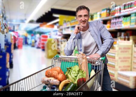 Lächelnder Supermarktkunde Mann schaut geradeaus und lehnt auf einen Warenkorb voller Lebensmittel im Supermarkt Gang Stockfoto