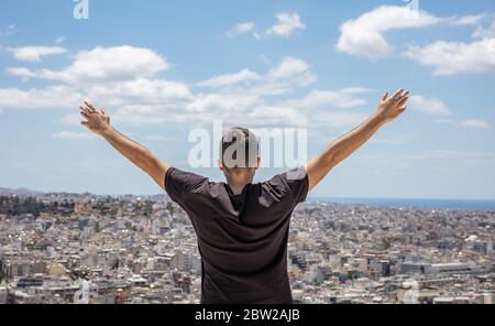 Mann zurück mit erhobenen Armen. Junge Erwachsene Rückansicht stehend allein mit offenen Händen Blick auf die Stadt unten, blauer Himmel Hintergrund. Gesunder Lebensstil, fre Stockfoto