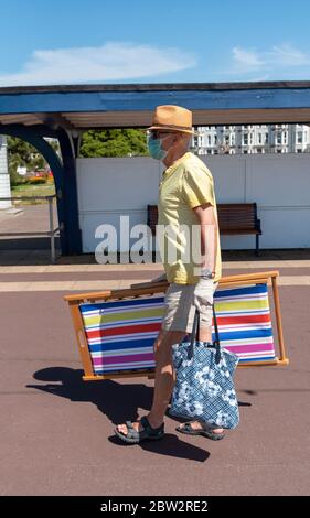 Southsea, Portsmouth, England, Großbritannien. Mai 2020. Mann, der während des Covid-19-Ausbruchs eine medizinische Maske und Handschuhe trägt, die einen Liegestuhl entlang der Promenade tragen. Stockfoto