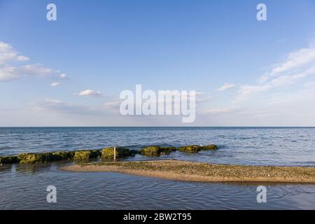 Ein kleines Sandband, das zu einem natürlichen Wellenbrecher führt. Mit blauem, sauberem Himmel und Wolken auf dem Hintergrund. Stockfoto