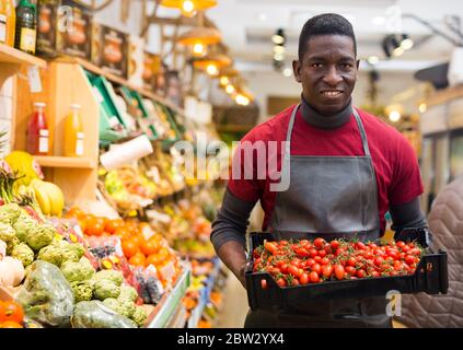 Fröhlicher afroamerikanischer Mann, der auf dem Bauernmarkt arbeitet und frische reife Kirschtomaten auf der Theke arrangiert Stockfoto