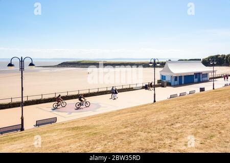 Ein paar Leute gehen und radeln entlang der Promenade am ruhigen Strand von Barry Island an einem sonnigen Frühlingsferiennachmittag während der 2020 Coronaviru Stockfoto