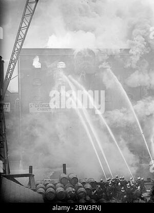 250 Feuerwehrmann Kampf london Reis Lager Flammen . Motoren von jedem der 60 Stationen im LCC wurden zu einem riesigen Feuer in St Georges ' s Wharf, ein Reislager von Carbutt and Company, Ltd. In Shad thames, Bermondsey, Ein Notruf wurde getätigt und jedes verfügbare Gerät wurde gesendet. Etwa 250 Männer waren auf den Ausbruch konzentriert. Feuerwehrleute trugen Gasmasken gegen den dichten Rauch aus dem Reis, und ein riesiger Drehtisch Wasserturm wurde in die Operation gebracht. Man befürchtete, dass eine Mauer einstürzen würde. Foto zeigt, Wasser aus den Schlauchleitungen Kaskadierung über das brennende Gebäude. Stockfoto