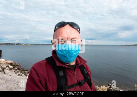 Am Flussufer steht ein Glatzkopf in einem roten Pullover mit medizinischer Schutzmaske und Sonnenbrille. Ein älterer Mann in einer Zeit der Selbstisolierung und q Stockfoto