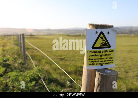 'Uneingezäunte Klippe mit Überhängen voraus' Postschild gelb und weiß mit grünem Feld im Hintergrund. Stockfoto