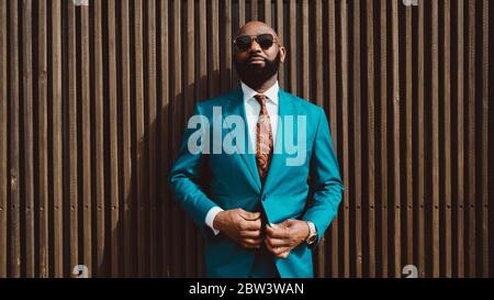 Ein schöner, reifer kahlbärtiger Afrikaner in Sonnenbrille und einem modischen blauen oder blaugrünen Kostüm mit Krawatte steht vor einer Wand Stockfoto