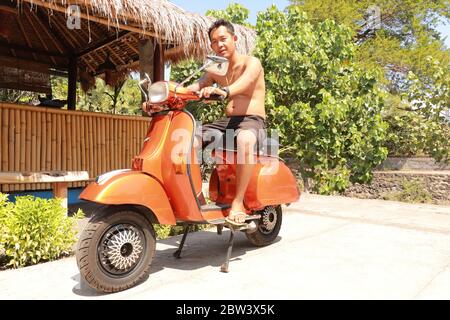 Bali, Indonesien, 1. Juli 2019. Youg Indonesier Mann sitzt auf orange Classic Vespa. Klassische Vespa In Orange. Ein alter oranger Vespa Super Motorroller Stockfoto