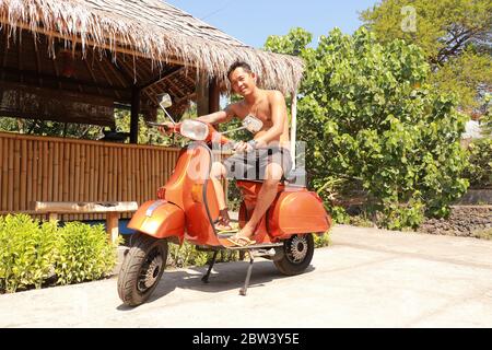 Bali, Indonesien, 1. Juli 2019. Youg Indonesier Mann sitzt auf orange Classic Vespa. Klassische Vespa In Orange. Ein alter oranger Vespa Super Motorroller Stockfoto