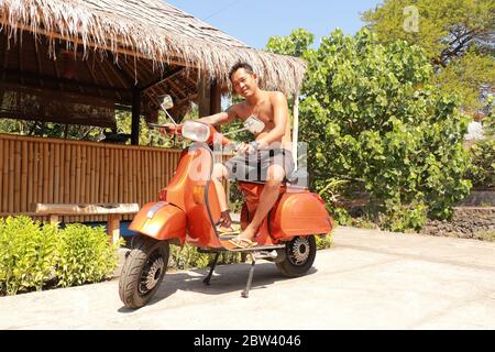 Bali, Indonesien, 1. Juli 2019. Youg Indonesier Mann sitzt auf orange Classic Vespa. Klassische Vespa In Orange. Ein alter oranger Vespa Super Motorroller Stockfoto