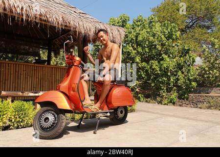 Bali, Indonesien, 1. Juli 2019. Youg Indonesier Mann sitzt auf orange Classic Vespa. Klassische Vespa In Orange. Ein alter oranger Vespa Super Motorroller Stockfoto