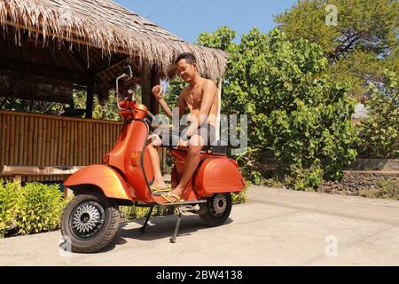 Bali, Indonesien, 1. Juli 2019. Youg Indonesier Mann sitzt auf orange Classic Vespa. Klassische Vespa In Orange. Ein alter oranger Vespa Super Motorroller Stockfoto