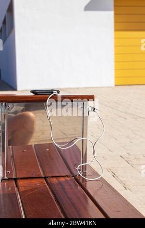 Solarbetriebenes USB-Ladegerät mit Handy auf Ladung und Bank draußen auf dem Fußweg in Playa San Juan, teneriffa, Kanarische Inseln, Spanien Stockfoto