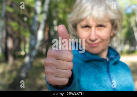 Eine Frau mittleren Alters lächelt und zeigt einen Daumen nach oben. Spaziergang im Park an einem hellen sonnigen Tag. Gute Laune. Stockfoto