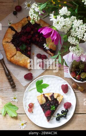 Süße Pie-Galette mit saftiger Beerenfüllung, Beeren und Wildblumen auf Holzhintergrund. Rustikaler Stil. Stockfoto