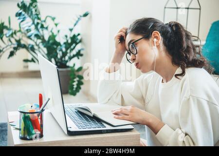 Müde kaukasische Frau, die am Computer studiert, während sie ein Buch liest und Kopfhörer trägt Stockfoto