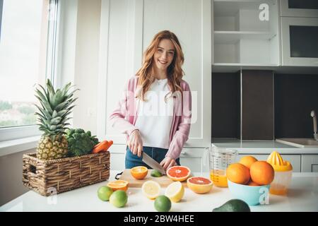 Kaukasische Frau mit roten Haaren und Sommersprossen lächelt in der Kamera, während sie schneiden Früchte und quetschen sie machen Saft aus Limette und Orange Stockfoto