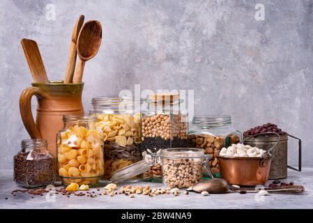 Verschiedene getrocknete Hülsenfrüchte und italienische Pasta in Glas und Metall Gläser. Graue Hintergrundstruktur. Stillleben Stockfoto