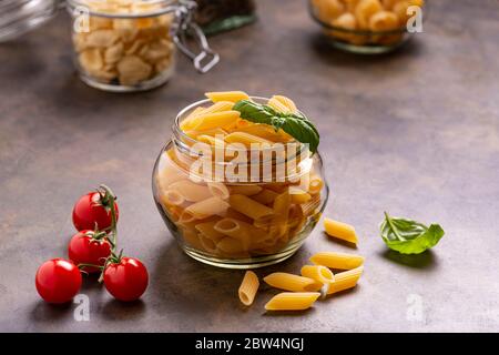 Im Vordergrund in einem transparenten Glas rohe italienische Pasta und frische Basilikumblätter. Im Hintergrund ein Bündel Kirschtomaten Stockfoto