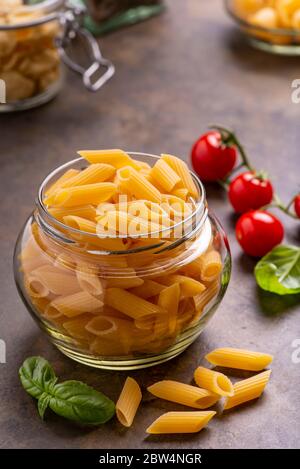 Im Vordergrund in einem transparenten Glasgefäß, rohe italienische Pasta, im Hintergrund ein Bündel Kirschtomaten und frische Basilikumblätter. Stockfoto