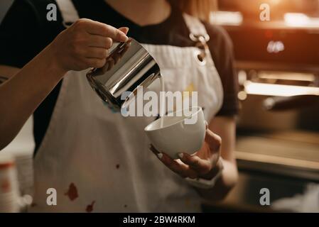 Eine Barista mit einem Becher Milch aus Metall bastelt in einem Café eine Latte-Kunst auf einen Kaffee. Ein Barista gießt Milch mit einem schmalen Bach, um zu vermeiden, dass der bricht Stockfoto