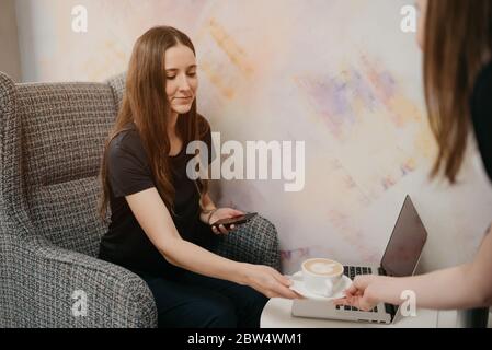 Eine Barista hält einem Mädchen in einem Café einen Latte. Ein Mädchen mit langen Haaren, das ferngesteuert an einem Laptop arbeitet, hält soziale Distanz, um eine Tasse zu ergreift Stockfoto