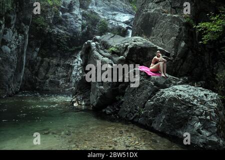 Eine griechische Frau, die auf einem Felsen nahe einem Wasserfall auf dem Fonias Fluß sitzt, auf der nordgriechischen Insel Samothraki, Nordägäis, Thrakien, Griechenland. Stockfoto