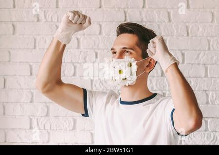 Portrait des fröhlichen männlichen Arztes in Blume Schutzmaske feiert Sieg gegen Coronavirus. Junge glückliche Mann in medizinischen Gesichtsmaske mit Gänseblümchen fl Stockfoto