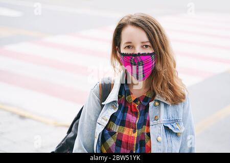 Junge Frau trägt eine karierte handgemachte Gesichtsmaske und buntes Hemd auf der Straße stehend. Stockfoto