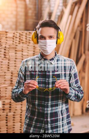 Portrait professioneller Zimmermann in medizinischer Maske auf dem Gesicht, Gehörschutz Kopfhörer tragen Schutzbrille am Sägewerk. Handwerker am Holz Stockfoto