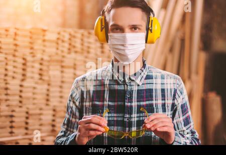 Portrait des jungen Zimmermanns in Gehörschutz Kopfhörer und medizinische Gesichtsmaske tragen Schutzbrille in der Werkstatt. Männlicher Handwerker im Sägewerk. Stockfoto