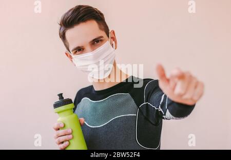 Selbstbewusster Sportmann in medizinischer Gesichtsmaske mit Zeigefinger und Musik hören. Glücklicher junger Mann Fitness-Trainer in Schutzmaske. Motiv Stockfoto