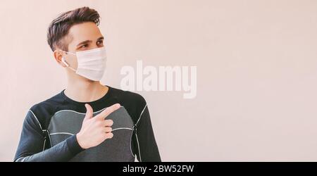 Junge zuversichtlich Sportmann in medizinischen Gesichtsmaske zeigt mit Finger auf Hintergrund mit Kopierraum. Happy Fitness Trainer in Schutzmaske zeigt c Stockfoto