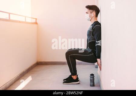Junger sportlicher Mann Fitness-Trainer in medizinischer Gesichtsmaske hockt gegen Wand auf Balkon. Selbstbewusste Sportmann in Schutzmaske tun Wand Bein sitzen exer Stockfoto